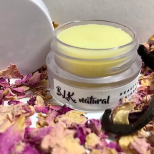 Pu&ograve; includere: Un piccolo barattolo trasparente con un balsamo per labbra giallo al suo interno. Il barattolo ha un'etichetta nera che recita "BLK natural". Il barattolo &egrave; circondato da petali di rosa secchi e una bacca di vaniglia.