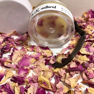 Pu&ograve; includere: Un barattolo di vetro trasparente con un coperchio bianco riempito di un balsamo giallo chiaro. Il barattolo ha un'etichetta nera su cui &egrave; scritto "BLK natural". Il barattolo &egrave; circondato da petali di rosa secchi e una bacca di vaniglia.