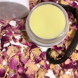 Pu&ograve; includere: Primo piano di un piccolo barattolo bianco pieno di un balsamo giallo pallido. Il barattolo &egrave; circondato da petali di rosa secchi e un baccello di vaniglia.