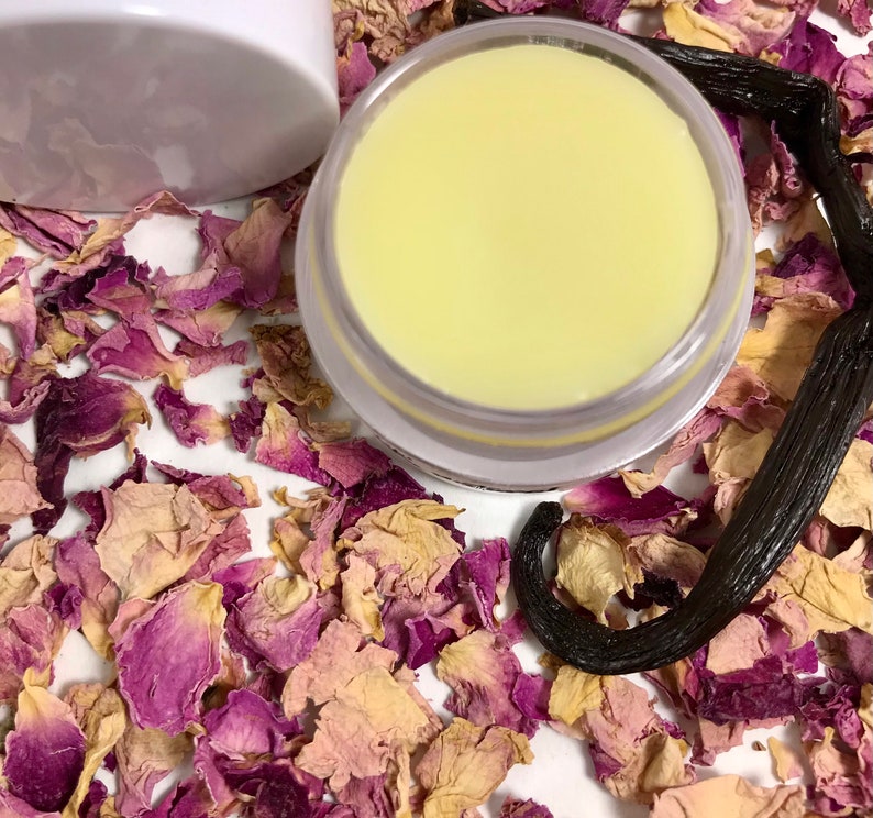 Pu&ograve; includere: Primo piano di un piccolo barattolo bianco pieno di un balsamo giallo pallido. Il barattolo &egrave; circondato da petali di rosa secchi e un baccello di vaniglia.