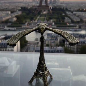 May include: A bronze Eiffel Tower figurine with a balancing eagle sculpture. The eagle has outstretched wings. The background features the actual Eiffel Tower and a cityscape at sunset.