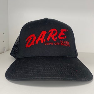 May include: Black trucker hat with the red text "D.A.R.E. TO KEEP COPS OFF DONUTS" embroidered on the front. The hat has a mesh back and a curved brim. The hat is sitting on a white surface.
