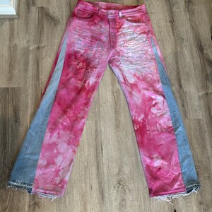 May include: A pair of denim jeans with pink and blue tie-dye patterns. The jeans have a distressed look with frayed edges.