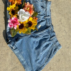 Puede incluir: Pantalones cortos de mezclilla azul claro con un diseño floral hecho de flores de tela. Las flores son amarillas, rosadas, blancas y naranjas.