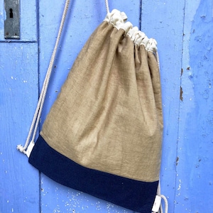 Op de afbeelding: Een bruin trekkoordrugzak met een marineblauw onderste paneel en witte trekkoordkoorden. De tas hangt tegen een verweerde blauwe houten deur. De bovenkant van de tas is crèmekleurig.