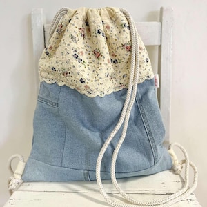 Può includere: Zaino con coulisse in denim con parte superiore in pizzo floreale. La borsa combina denim blu chiaro con una sezione in pizzo color crema ornata da un motivo floreale rosso, blu e verde. Ha cinghie di corda bianche.