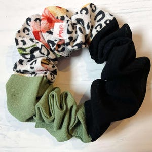May include: A colourful scrunchie with sections of floral, leopard print, olive green, and black fabric. The scrunchie is circular and made of soft fabric. A small tag with text is visible.