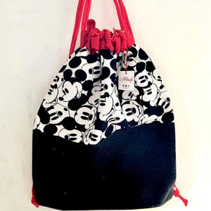 Könnte beinhalten: Ein Kordelzugrucksack mit einem schwarz-weißen Mickey Mouse-Muster auf dem oberen Teil und einem einfarbigen schwarzen unteren Teil. Die Tasche hat einen roten Kordelzug und einen roten oberen Teil. Ein kleines Etikett ist an der Tasche befestigt.