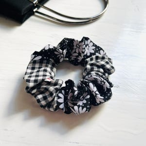 Puede incluir: Una goma para el pelo en blanco y negro con una mezcla de estampados, incluyendo cuadros de vichy y diseños florales. El accesorio para el cabello está hecho de tela. Un asa de bolso negro es visible en el fondo.