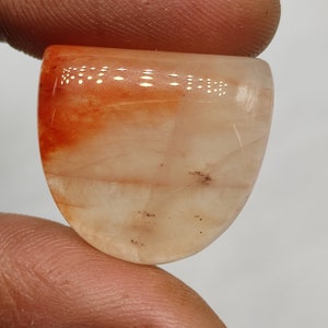 May include: A cabochon cut, translucent, light orange and white gemstone with brown inclusions. The gemstone is oval shaped and has a smooth, polished surface.