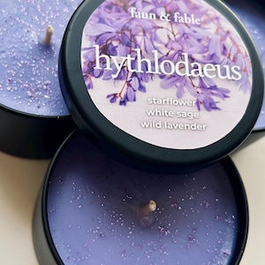 May include: Three small, round, black metal tins of scented candles are shown. The candles are a light purple colour with glitter and a wooden wick. The lids feature a floral design and the text "faun & fable" and "hythlodaeus", with scent notes listed below.