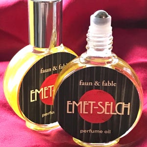May include: Two small, round perfume oil bottles with a golden liquid. Each bottle has a black label with the text "faun & fable" and "EMET-SELCH perfume oil" in white. One bottle has a silver cap, the other a rollerball.