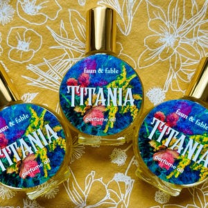 May include: Three perfume oil bottles with gold caps and labels that read "faun & fable TITANIA perfume oil." The labels have a blue background with floral and butterfly designs. The bottles are on a yellow floral patterned surface.