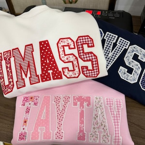 May include: Three sweatshirts in white, navy, and pink. The white sweatshirt has "UMASS" in red letters with floral and polka dot patterns. The navy sweatshirt has "VS" in white letters with a floral pattern. The pink sweatshirt has "TAYTAY" in pink letters with floral patterns.
