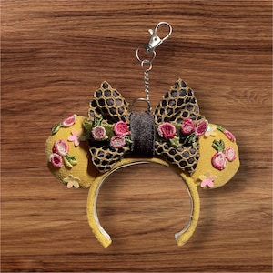 Op de afbeelding: Gele stoffen hoofdband met bevestigde oren en een strik. De oren zijn versierd met roze bloemenborduurwerk en kleine roze bloemen. De strik is zwart met een honingraatpatroon en een bruin midden. Een zilveren sleutelhanger is bevestigd.