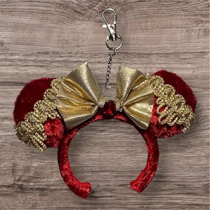 May include: Red velvet mouse ear headband with a gold bow and trim. A red jewel is centered on the bow. The headband has a silver keychain attached to the top. The ears are trimmed with gold decorative trim.