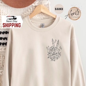 May include: Sand-colored sweatshirt with a black skeleton hand making a peace sign, surrounded by flowers and sparkles. The sweatshirt is hanging on a wooden hanger. The words "Next Day Shipping" are visible on a sticker.