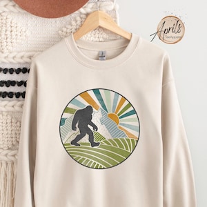May include: A beige sweatshirt with a retro-style graphic of a Bigfoot walking in front of mountains with a sun rising behind them. The graphic is in a circle with a green, blue, and orange color scheme.