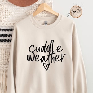 May include: A beige sweatshirt with the text "Cuddle Weather" in black script with a small heart underneath.