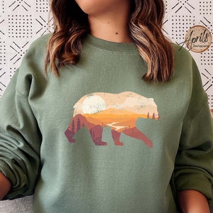 May include: A green sweatshirt with a graphic print of a bear standing in front of a mountain range with a sunset in the background.