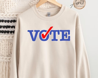May include: A beige sweatshirt with the word "VOTE" in blue letters with a red checkmark over the "O".