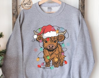 May include: A gray sweatshirt with a brown Highland cow wearing a Santa hat and surrounded by Christmas lights.