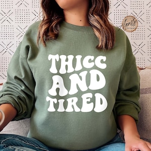 May include: A green sweatshirt with the text "THICC AND TIRED" in white, retro-style lettering. The model is wearing the sweatshirt and sitting on a couch.