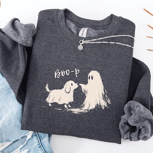 May include: A dark charcoal gray crewneck sweatshirt featuring a graphic of a small dog and a ghost with the text "BOO-p" in white. The sweatshirt is paired with light blue distressed denim jeans and a silver-toned chain necklace.
