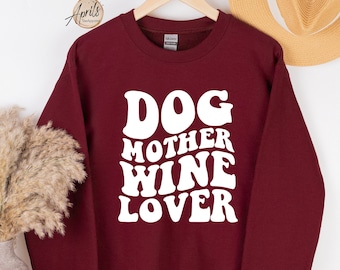 May include: A maroon sweatshirt with the words "DOG MOTHER WINE LOVER" in white, retro-style lettering. The sweatshirt is hanging on a wooden hanger. A straw hat and a vase with dried plants are in the background.