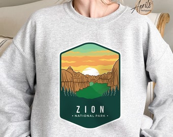 May include: A gray sweatshirt with a graphic design of a sunset over a mountain range with a lake in the foreground. The graphic is in a green, orange, and brown color scheme and has the text "Zion National Park" in white.