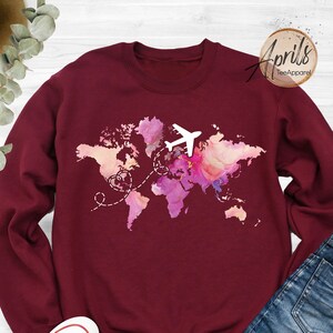 May include: Burgundy sweatshirt with a watercolor world map design and a white airplane flying over it. The map has pink, purple, and blue hues. The text "Aprils Tee Apparel" is in the top right corner.