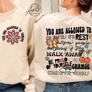 May include: Beige sweatshirt with the text "YOU ARE ALLOWED TO THRIVE" and a flower graphic on the front. The back features the text "YOU ARE ALLOWED TO" followed by inspirational phrases and graphics. A casual, comfortable top.