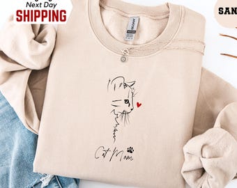 May include: A sand-coloured sweatshirt featuring a minimalist cat sketch and the words "Cat Mom" in black. A small red heart is next to the cat's face. The sweatshirt is paired with blue jeans.