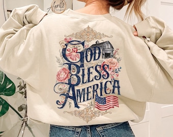 May include: A cream-coloured sweatshirt with a graphic print of a barn, flowers, and the text "God Bless America" in blue and red lettering. The graphic also includes a small American flag.