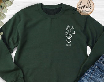Puede incluir: Una sudadera verde oscuro con un gráfico blanco de un signo zodiacal de Tauro con una luna creciente y flores. El texto "Taurus" está debajo del gráfico.