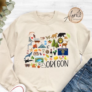 May include: A beige sweatshirt with a graphic design of the state of Oregon. The design features a heart with the word "OREGON" inside and is surrounded by various icons representing things associated with the state, such as a pine tree, a beaver, a coffee cup, and a wine bottle. The sweatshirt is laid flat on a white surface with a pair of white sneakers and blue jeans.