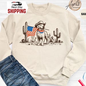 Pu&ograve; includere: Felpa color sabbia con una grafica di un cane con cappello da cowboy e bandiera americana. Il cane &egrave; in una scena desertica con cactus. La felpa &egrave; mostrata con jeans blu e scarpe da ginnastica bianche.