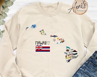 May include: A beige sweatshirt with a graphic of the Hawaiian islands, featuring a variety of icons representing the state's culture and nature. The text "HAWAII ♥" and the Hawaiian flag are also included in the design.