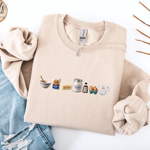 May include: A beige crewneck sweatshirt featuring a graphic print of baking ingredients including a mixing bowl, a bag of flour, a stick of butter, a jar of sugar, a bottle of vanilla extract, a carton of eggs, and a glass measuring cup.