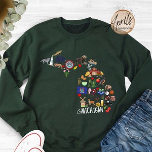 May include: A dark green sweatshirt with a graphic of the state of Michigan. The graphic is made up of various icons representing things associated with Michigan, such as a lighthouse, a cherry, a windmill, and a baseball. The text "MICHIGAN" is written in white below the graphic.