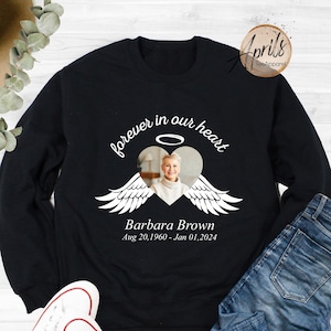 May include: Black sweatshirt with white text and a photo of a woman in a heart with angel wings. The text reads "forever in our heart" and "Barbara Brown Aug 20, 1960 - Jan 01, 2024".