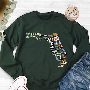 May include: A dark green sweatshirt with a graphic of the state of Florida. The graphic includes various icons representing Florida, such as palm trees, oranges, and a lighthouse. The text "The Sunshine State" and "Florida" are also included in the graphic.