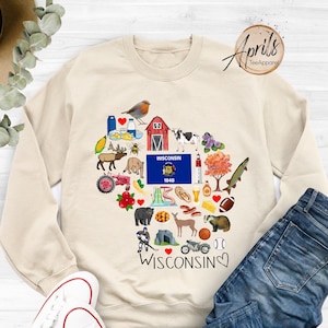 May include: A cream-colored sweatshirt with a heart-shaped design featuring various icons representing Wisconsin. The design includes the state flag, a red barn, a cow, a deer, a bear, a cheese wedge, a bratwurst, a football, and the text "WISCONSIN 1848". The sweatshirt also has the text "WISCONSIN" with a heart symbol.