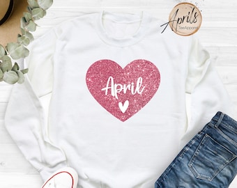 May include: White sweatshirt with a pink glitter heart that says "April" on it. The sweatshirt is laying on a white surface with a pair of white sneakers and blue jeans.