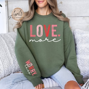 May include: A green sweatshirt with the text "LOVE more" in pink and red lettering. The left sleeve has the text "WORRY less" in pink and red lettering.