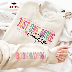 May include: A sand-colored sweatshirt with the text "JUST ONE MORE chapter" in colorful letters. The sleeve displays the word "BOOKWORM" in a rainbow of colors. A heart graphic is next to the word "chapter".