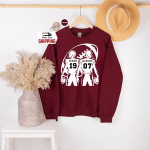May include: A maroon sweatshirt with a white graphic of two football players inside a football. The graphic includes the names "MARK" and "JACKSON" with the numbers "19" and "07". A straw hat hangs in the background.