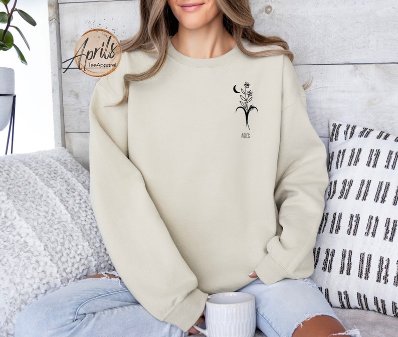 Op de afbeelding: Een licht beige sweatshirt met een zwarte afbeelding van een bloem en een wassende maan met het sterrenbeeld Ram en het woord "Ram" eronder.