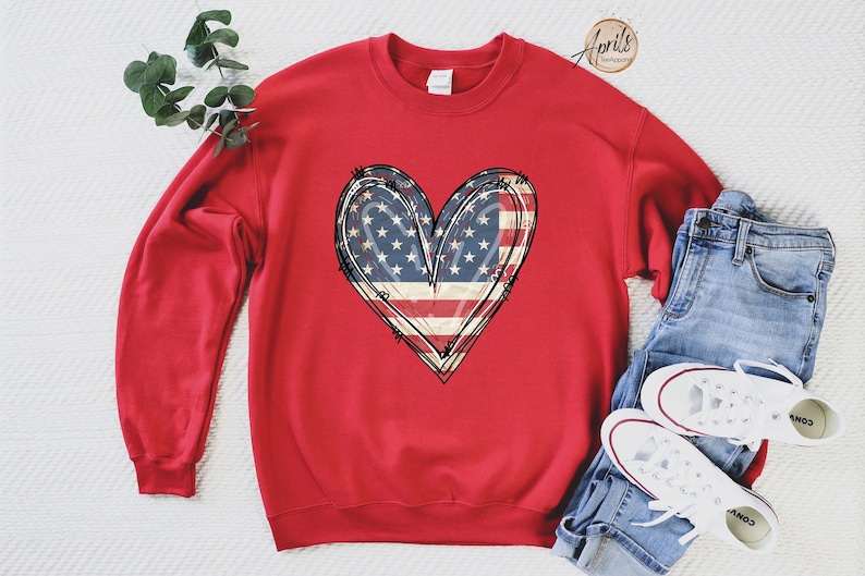 Puede incluir: Sudadera roja con un dise&ntilde;o de coraz&oacute;n de bandera estadounidense blanca y azul. La sudadera est&aacute; colocada sobre una superficie blanca con un par de pantalones vaqueros azules y zapatillas blancas.
