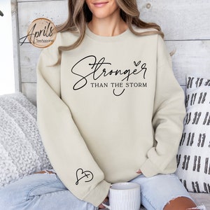 May include: A beige sweatshirt with the text "Stronger than the storm" and a small heart design. The sweatshirt has a small heart with a cross inside on the sleeve.